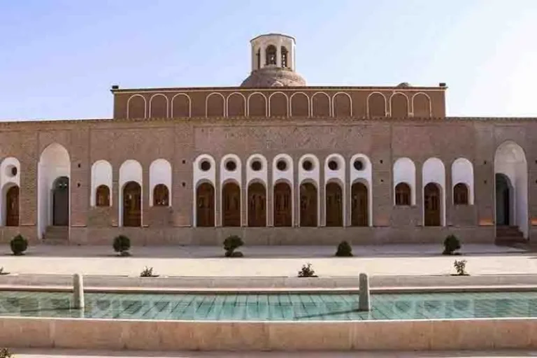 نمای حیاط یا ورودی خانه حاج علی‌آقا با معماری سنتی، پنجره‌های چوبی و تزئینات گچ/آجر، در نور روز.